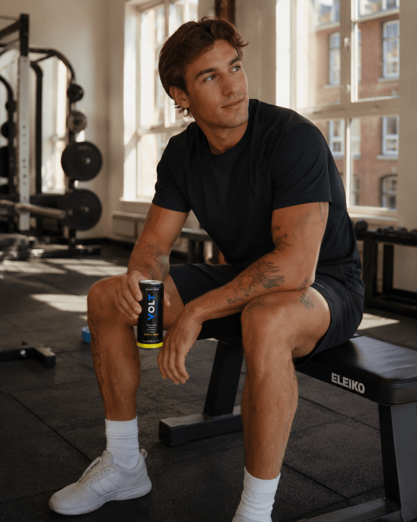 Photorealistic person holding VOLT Arctic Mint energy drink can at premium Amsterdam fitness studio after workout in quality gym wear with natural window light and clean gym equipment in background