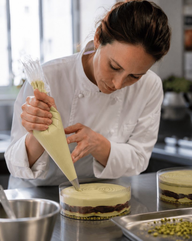 Photorealistic Maison Florine pastry chef Camille Beaumont in white coat piping precise pistachio mousse layer into clear acetate entremets mould on stainless steel bench with natural laboratoire light