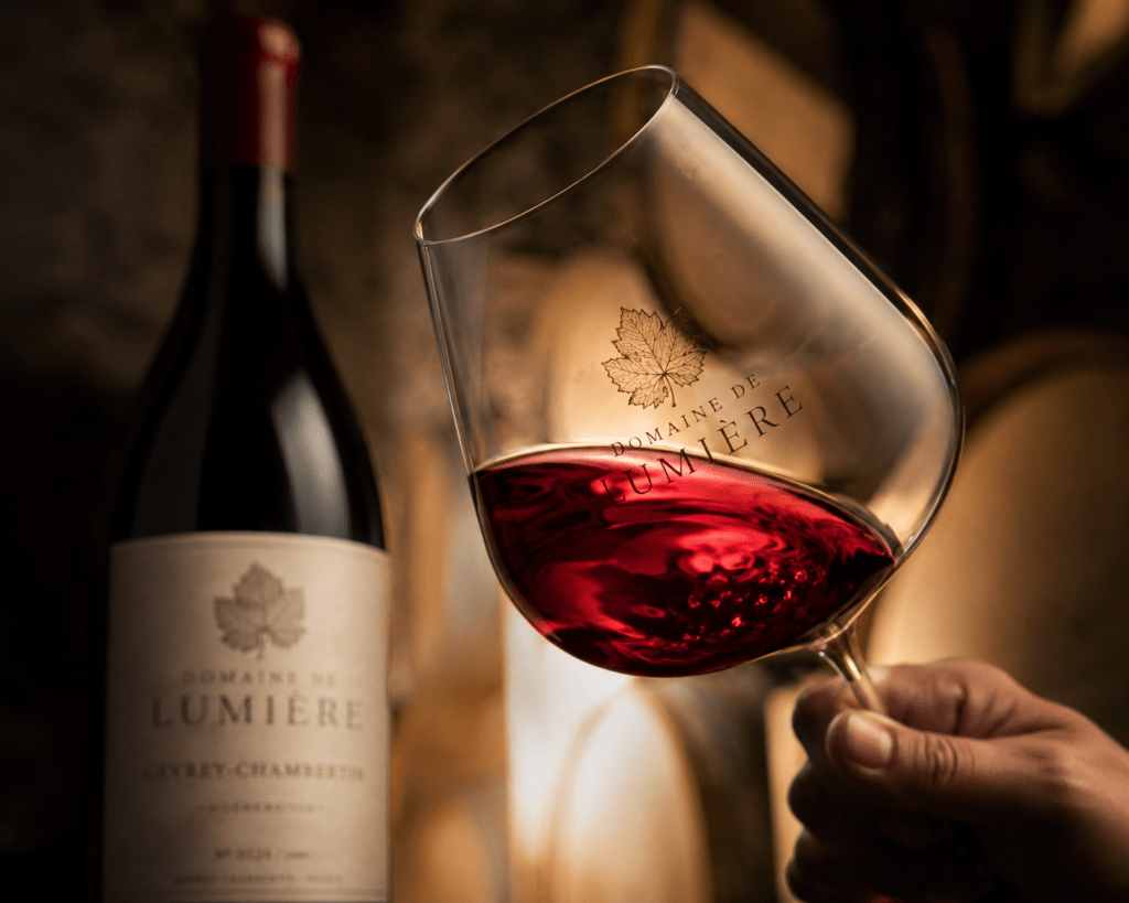 Photorealistic Domaine de Lumière Pinot Noir poured into traditional Burgundy tasting glass showing translucent garnet ruby colour backlit against blurred limestone cellar barrel background