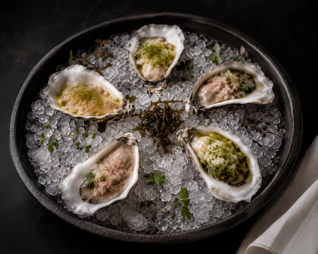 Six Pacific oysters on crushed ice on slate dish each with different mignonette garnishes and micro herbs