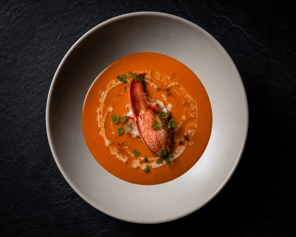 Fine dining lobster bisque in cream porcelain bowl with lobster claw, truffle cream oil and micro chervil on dark slate